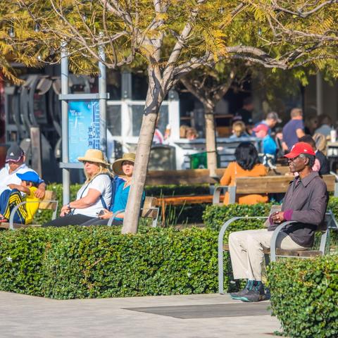 People sitting on benches under the trees and sun at Broadway Plaza at the Port of San Diego
