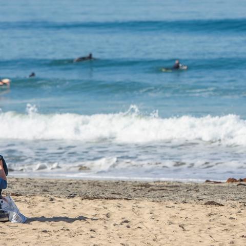 Man sitting on the sandy shore in front of surfers paddling over the waves at Portwood Pier Plaza at the Port of San Diego