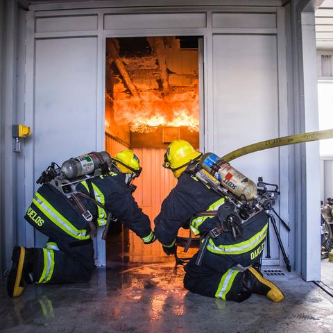 two Port of San Diego Harbor Police Firefighters training in front of a door with fire raging 