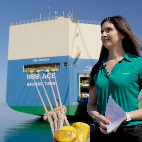 a woman with long dark hair, wearing a green shirt, stands on a dock in front of a cargo ship names Iris Ace