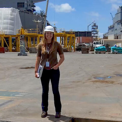 a woman wearing jeans, brown t-shirt, white hard-hat and carrying a clipboard, smiles at the camera. She is in front of large gray ships