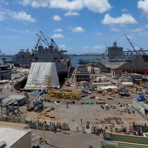 an aerial view of 3 large, gray ships in dry-dock. the shipyard is in the foreground